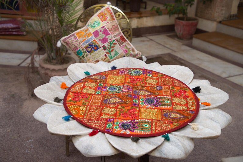 May include: A round, orange-toned fabric table mat featuring a patchwork design with intricate embroidery, metallic thread accents, and small mirrors. The mat is finished with a dark red border and several small, multi-coloured fabric tassels.