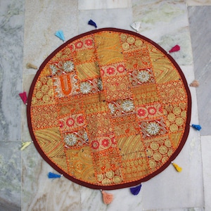May include: Circular patchwork floor cushion in orange, gold, and red hues. The cushion has detailed embroidery and is edged with a dark red border and colourful tassels. A decorative accent for any room, approximately 75 cm in diameter.