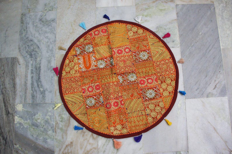 May include: Circular patchwork floor cushion in orange, gold, and red hues. The cushion has detailed embroidery and is edged with a dark red border and colourful tassels. A decorative accent for any room, approximately 75 cm in diameter.