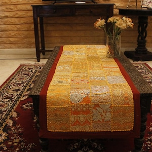 Może przedstawiać: Prostokątny stolik kawowy z bieżnikiem w stylu patchwork. Bieżnik jest głównie żółty z czerwonymi obramowaniami i mieszanką wzorzystych kwadratów. W tle wzorzysty dywan i drewniane krzesło.