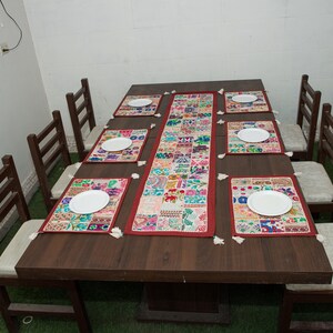 Puede incluir: Una mesa de comedor rectangular de madera marr&oacute;n oscuro con seis sillas con cojines blancos. La mesa est&aacute; decorada con un camino de mesa largo de tela de retazos multicolor y seis manteles individuales rectangulares a juego, cada uno con un plato blanco.