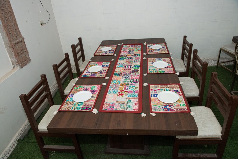 Puede incluir: Una mesa de comedor rectangular de madera marr&oacute;n oscuro con seis sillas con cojines blancos. La mesa est&aacute; decorada con un camino de mesa largo de tela de retazos multicolor y seis manteles individuales rectangulares a juego, cada uno con un plato blanco.