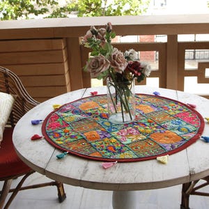 May include: A round white wooden table with two matching rattan chairs, each featuring a red seat cushion and a white patterned throw pillow. A circular, multicoloured embroidered fabric mat with small tassels sits under a glass vase of roses.