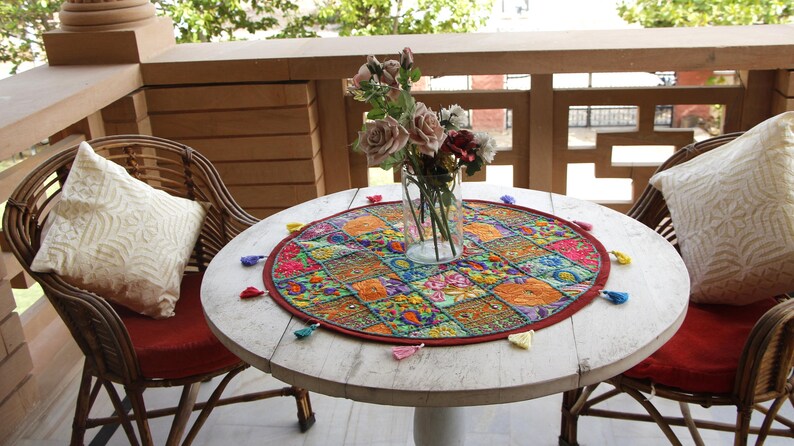 May include: A round white wooden table with two matching rattan chairs, each featuring a red seat cushion and a white patterned throw pillow. A circular, multicoloured embroidered fabric mat with small tassels sits under a glass vase of roses.