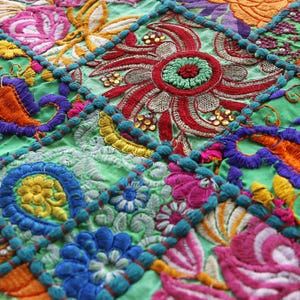 K&ouml;nnte beinhalten: Nahaufnahme eines farbenfrohen, bestickten Textils mit Patchwork-Design. Der Stoff weist lebendige Blumen- und Paisley-Muster in Orange-, Lila-, Rosa- und Gr&uuml;nt&ouml;nen auf. Die Stickerei ist detailliert und aufwendig.