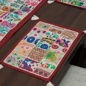 Puede incluir: Un mantel individual rectangular que presenta un mosaico de cuadrados de tela bordados en colores vibrantes, incluidos rojo, azul, verde y amarillo. El mantel est&aacute; bordeado con tela roja oscura s&oacute;lida y rematado con peque&ntilde;as borlas blancas en cada esquina.