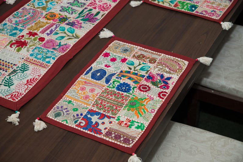 Puede incluir: Un mantel individual rectangular que presenta un mosaico de cuadrados de tela bordados en colores vibrantes, incluidos rojo, azul, verde y amarillo. El mantel est&aacute; bordeado con tela roja oscura s&oacute;lida y rematado con peque&ntilde;as borlas blancas en cada esquina.