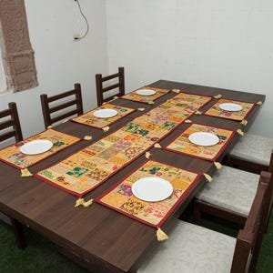 Puede incluir: Una mesa de comedor de madera marr&oacute;n oscuro con seis sillas que tienen cojines de color claro. La mesa est&aacute; decorada con un camino de mesa rectangular de patchwork y seis manteles individuales rectangulares a juego, cada uno con un plato blanco.