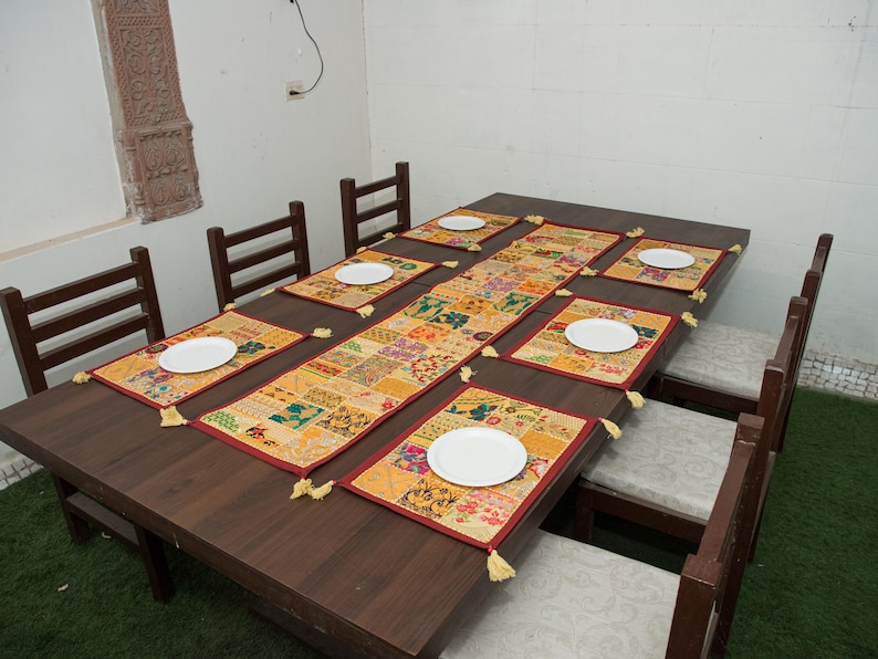 Puede incluir: Una mesa de comedor de madera marr&oacute;n oscuro con seis sillas que tienen cojines de color claro. La mesa est&aacute; decorada con un camino de mesa rectangular de patchwork y seis manteles individuales rectangulares a juego, cada uno con un plato blanco.