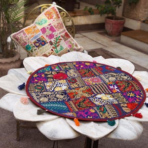 May include: A round, multi-coloured patchwork fabric mat featuring intricate embroidery and gold-toned thread accents. The mat has a dark red border and is decorated with small, colourful fabric tassels attached around its circular edge.