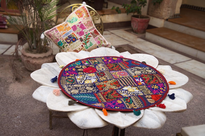 May include: A round, multi-coloured patchwork fabric mat featuring intricate embroidery and gold-toned thread accents. The mat has a dark red border and is decorated with small, colourful fabric tassels attached around its circular edge.