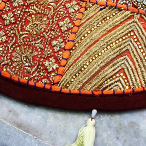 May include: A round floor cushion cover featuring a patchwork design of red and orange fabric panels. The surface is heavily embellished with intricate gold metallic embroidery, sequins, and beadwork. A dark red border with light tassels.