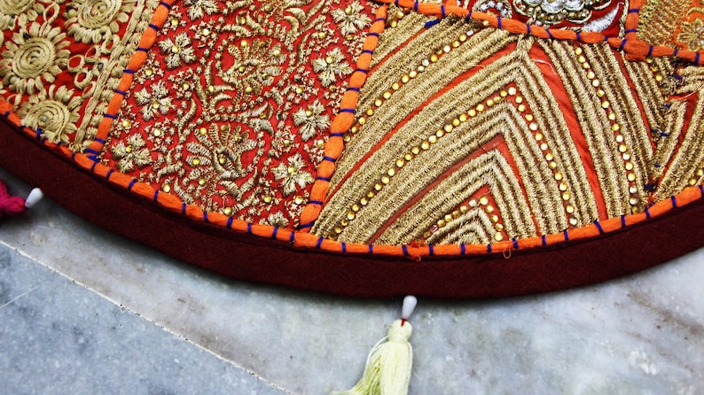 May include: A round floor cushion cover featuring a patchwork design of red and orange fabric panels. The surface is heavily embellished with intricate gold metallic embroidery, sequins, and beadwork. A dark red border with light tassels.