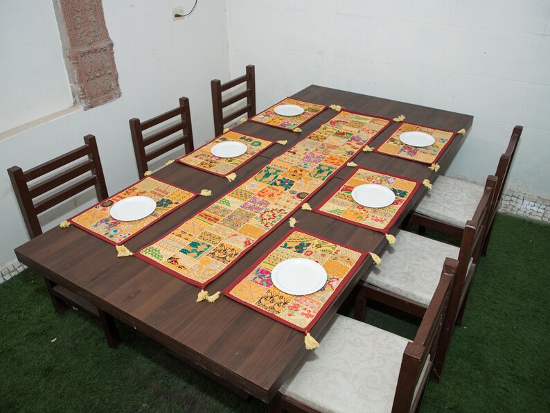 Puede incluir: Una mesa de comedor rectangular de madera marr&oacute;n oscuro con seis sillas con cojines claros. La mesa est&aacute; decorada con un camino de mesa de patchwork largo y seis manteles individuales rectangulares a juego, cada uno con un plato blanco.