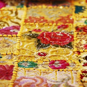 Puede incluir: Un camino de mesa rectangular con un dise&ntilde;o de patchwork de cuadrados de tela amarilla. Cada cuadrado est&aacute; decorado con intrincados bordados florales en tonos rojos, rosas, azules y verdes, acentuados con peque&ntilde;as lentejuelas y cuentas reflectantes.