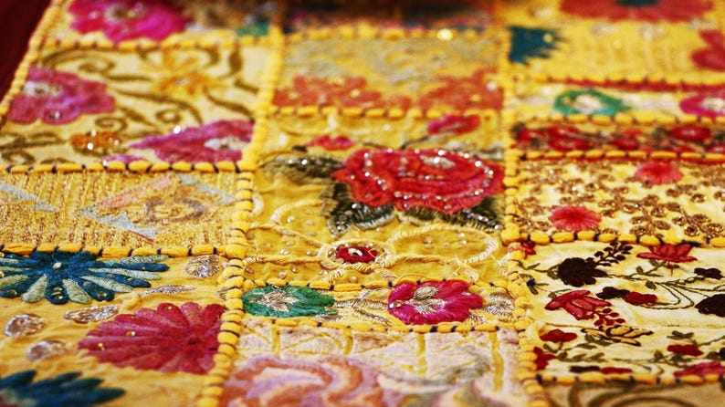 Puede incluir: Un camino de mesa rectangular con un dise&ntilde;o de patchwork de cuadrados de tela amarilla. Cada cuadrado est&aacute; decorado con intrincados bordados florales en tonos rojos, rosas, azules y verdes, acentuados con peque&ntilde;as lentejuelas y cuentas reflectantes.
