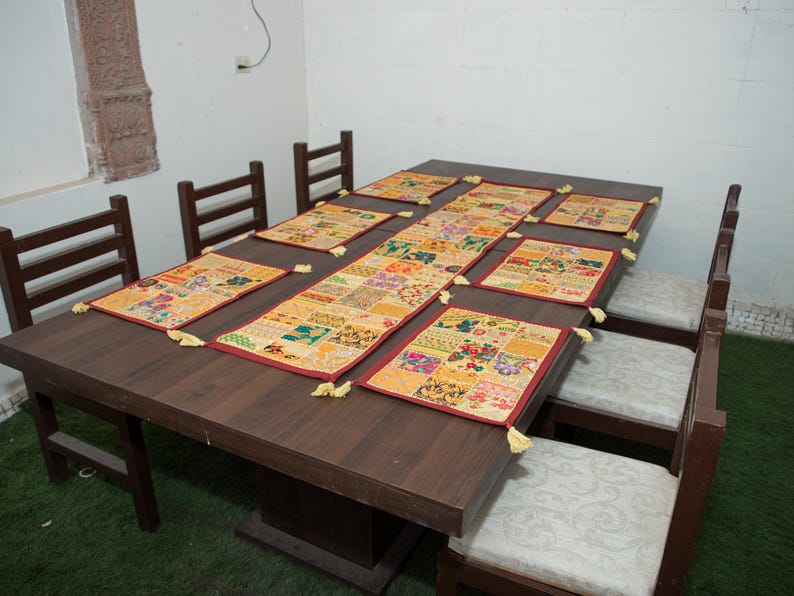 Puede incluir: Un juego de mesa de comedor rectangular que incluye un camino de mesa central largo y seis manteles individuales rectangulares a juego. Cada pieza est&aacute; hecha de un mosaico de cuadrados de tela multicolor con bordados y borlas amarillas.
