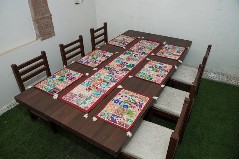 Puede incluir: Una mesa de comedor de madera marr&oacute;n oscuro con seis sillas a juego con cojines de colores claros. La mesa est&aacute; decorada con un camino de mesa rectangular de patchwork y seis manteles individuales rectangulares a juego, todos con borlas blancas.