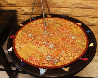 Puede incluir: Un mantel individual redondo de tela en tonos naranja con un diseño de patchwork de varios patrones florales y geométricos intrincados en oro, rojo y amarillo. El tapete circular tiene un borde rojo oscuro y pequeñas borlas de colores.