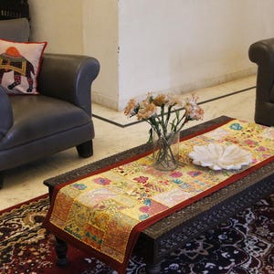 May include: Living room scene with a dark brown coffee table, a patterned runner, and a vase of flowers. A gray armchair with a decorative pillow and a dark brown sofa with a green pillow are also present. The room has a rug with a red and black pattern.