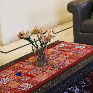 Könnte beinhalten: Ein dunkler Holztisch mit einem lebendigen roten Patchwork-Läufer. Eine klare Glasvase enthält getrocknete Blumen. Zwei dunkle Ledersessel flankieren den Tisch. Ein grünes Kissen liegt auf dem Sofa.