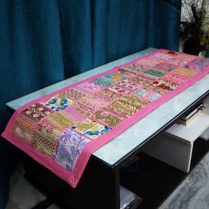 Könnte beinhalten: Ein rechteckiger Patchwork-Tischläufer mit rosa Rand. Der Läufer besteht aus verschiedenen Stoffquadraten in Rosa-, Gold- und Lilatönen. Der Läufer liegt auf einem schwarz-weißen Tisch.