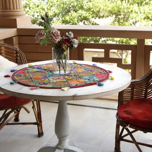 Pu&ograve; includere: Un tavolino rotondo bianco con un colorato tappetino fantasia e un vaso di fiori. Due sedie in vimini con cuscini rossi e cuscini color crema sono ai lati del tavolo. La scena &egrave; ambientata su un balcone.