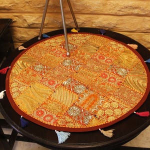 May include: A circular table mat with a patchwork design in gold, orange, and red hues. It features a dark red border and small tassels in various colours. The mat is placed on a dark brown table, approximately 75 cm in diameter.