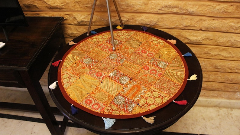 May include: A circular table mat with a patchwork design in gold, orange, and red hues. It features a dark red border and small tassels in various colours. The mat is placed on a dark brown table, approximately 75 cm in diameter.