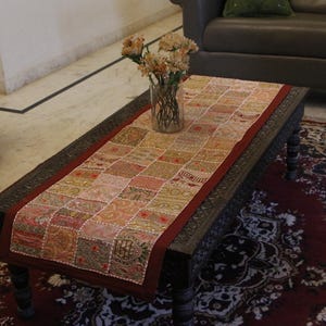 May include: A rectangular coffee table with a patchwork runner. The runner features a pattern of small, colorful squares in shades of pink, gold, and red. A vase of flowers sits on the table. The table is set on a patterned rug.