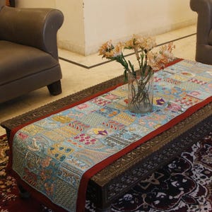 May include: A carved wooden coffee table with a patchwork runner in shades of blue, red, and yellow. A glass vase holds flowers on the table. The table is in a room with a brown leather armchair. The runner is approximately 150 cm long.