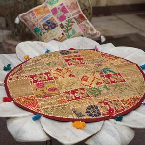 May include: A round, multi-coloured patchwork floor cushion featuring intricate gold, red, and blue fabric squares with floral and geometric embroidery. The circular cushion is finished with a dark red border and small, colourful fabric tassels.