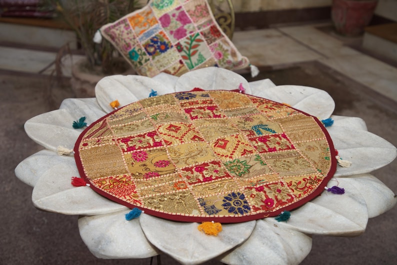 May include: A round, multi-coloured patchwork floor cushion featuring intricate gold, red, and blue fabric squares with floral and geometric embroidery. The circular cushion is finished with a dark red border and small, colourful fabric tassels.