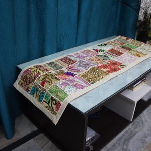 Könnte beinhalten: Ein farbenfroher Patchwork-Tischläufer mit cremefarbenem Rand liegt auf einem rechteckigen Tisch. Der Läufer zeigt eine Vielzahl von gemusterten Quadraten in Grün, Rot, Rosa und Gold. Der Tisch ist schwarz mit einer Glasplatte, der Läufer ist etwa 120 cm lang.