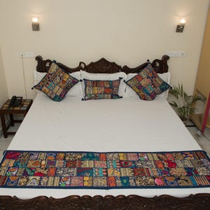 May include: A bed set featuring a white bedspread and a long rectangular bed runner decorated with a colorful patchwork pattern of various embroidered fabrics. Two square pillows and one rectangular pillow match the runner's patchwork design.