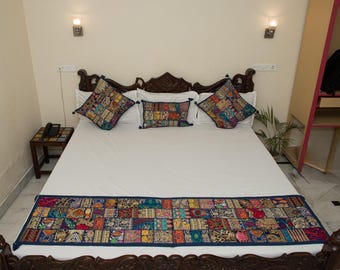 May include: A bed set featuring a white bedspread and a long rectangular bed runner decorated with a colourful patchwork pattern of various embroidered fabrics. Two square pillows and one rectangular pillow match the runner's patchwork design.