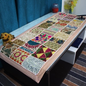 Pode incluir: Um caminho de mesa retangular com um design de patchwork de vários quadrados de tecido bordados em cores vibrantes, incluindo rosa, verde, dourado e azul. O caminho tem uma borda bege clara e é decorado com padrões florais complexos.