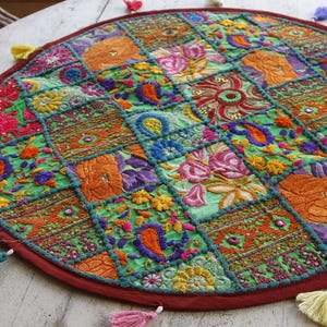 May include: A round, multi-coloured fabric table mat featuring a patchwork design of embroidered floral and paisley patterns in shades of green, orange, pink, and blue. The mat is bordered by a dark red trim and accented with small colourful tassels.