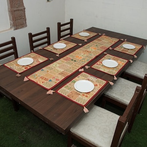 Könnte beinhalten: Ein dunkelbrauner, rechteckiger Esstisch aus Holz mit acht passenden Stühlen, die helle Sitzpolster haben. Der Tisch ist mit einem langen Patchwork-Tischläufer und sechs passenden rechteckigen Platzsets gedeckt, auf denen jeweils ein weißer Teller steht.
