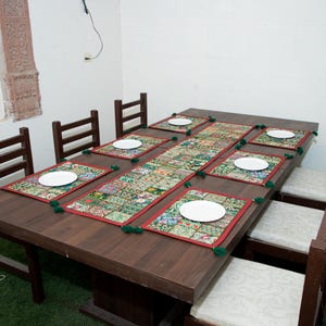 Puede incluir: Una mesa de comedor rectangular de madera marr&oacute;n oscuro con ocho sillas a juego con cojines blancos. La mesa est&aacute; decorada con un camino de mesa largo de tela de retazos multicolor y seis manteles individuales rectangulares a juego con platos blancos.