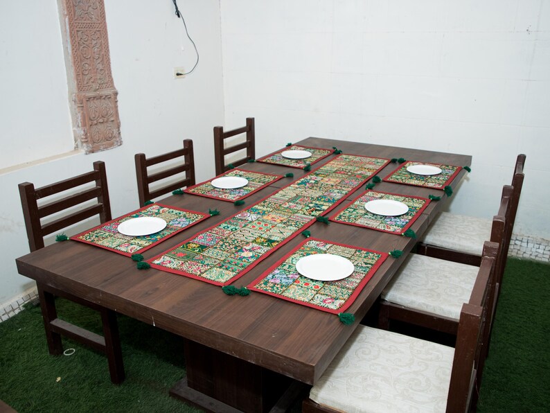 Puede incluir: Una mesa de comedor rectangular de madera marr&oacute;n oscuro con ocho sillas a juego con cojines blancos. La mesa est&aacute; decorada con un camino de mesa largo de tela de retazos multicolor y seis manteles individuales rectangulares a juego con platos blancos.