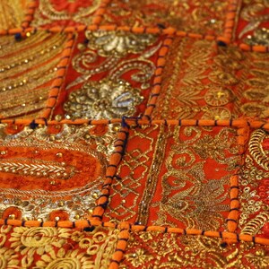 May include: Close-up of a patchwork textile with detailed embroidery. The design features a vibrant mix of orange, gold, and silver colours. The fabric is divided into square and rectangular sections, each adorned with detailed patterns and beadwork.