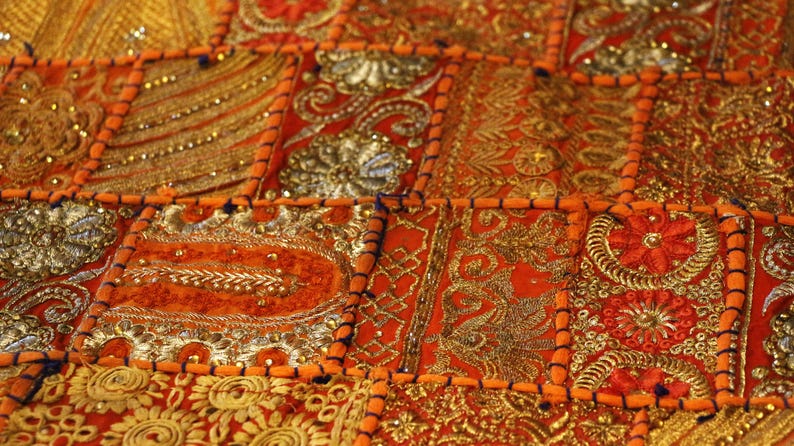 May include: Close-up of a patchwork textile with detailed embroidery. The design features a vibrant mix of orange, gold, and silver colours. The fabric is divided into square and rectangular sections, each adorned with detailed patterns and beadwork.