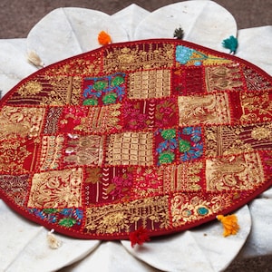 May include: A round, red fabric mat featuring a patchwork design of embroidered gold, green, and blue floral and geometric patterns. The edge of the mat is decorated with small, multi-coloured tassels in shades of yellow, pink, blue, and black.