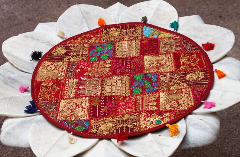 May include: A round, red fabric mat featuring a patchwork design of embroidered gold, green, and blue floral and geometric patterns. The edge of the mat is decorated with small, multi-coloured tassels in shades of yellow, pink, blue, and black.