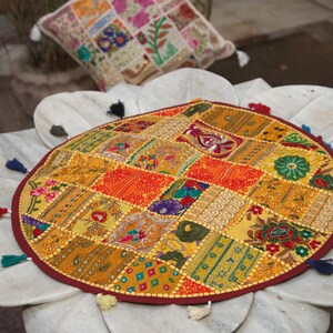 May include: A round, multi-coloured patchwork floor cushion featuring a variety of embroidered fabric squares in shades of yellow, orange, red, and green. The cushion is finished with a dark red border and small, multi-coloured tassels along the edge.