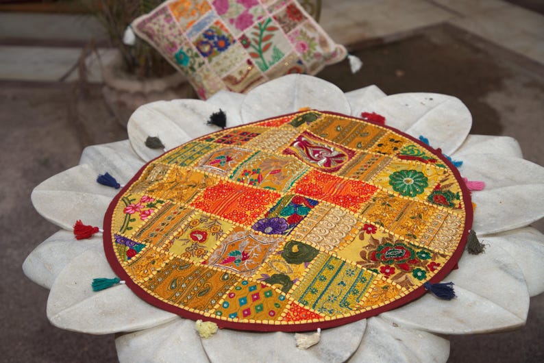 May include: A round, multi-coloured patchwork floor cushion featuring a variety of embroidered fabric squares in shades of yellow, orange, red, and green. The cushion is finished with a dark red border and small, multi-coloured tassels along the edge.