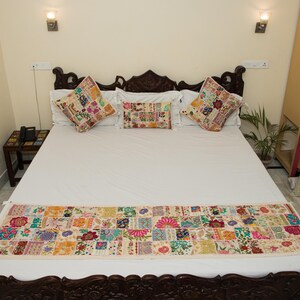 May include: A bed set featuring a white bedspread, a long rectangular bed runner, and three throw pillows. Each piece is decorated with a colourful patchwork design of embroidered floral and geometric patterns in various bright thread colours.