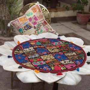 May include: A round, multi-coloured fabric mat featuring a patchwork of embroidered floral and geometric patterns in shades of red, blue, and gold, finished with a dark red border and small, multi-coloured tassels attached around the outer edge.