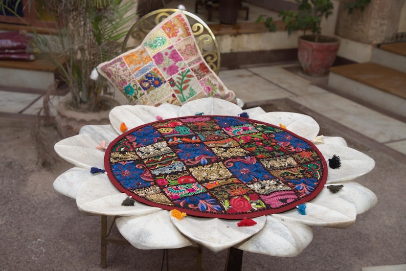May include: A round, multi-coloured fabric mat featuring a patchwork of embroidered floral and geometric patterns in shades of red, blue, and gold, finished with a dark red border and small, multi-coloured tassels attached around the outer edge.